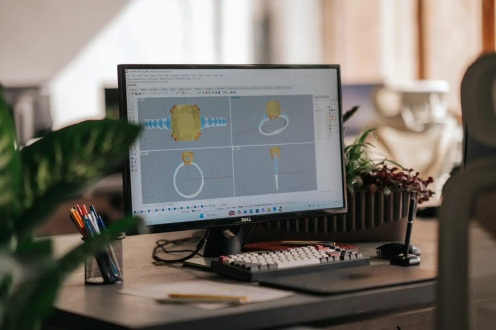 A computer monitor displaying a 3D ring design in CAD software on a desk surrounded by plants.