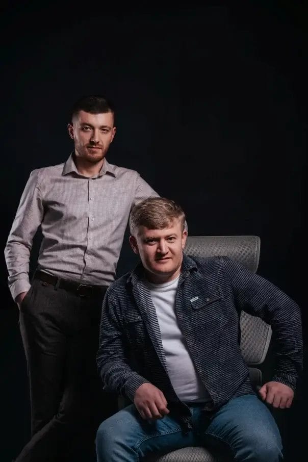 Two men posing against a dark background, one standing and one seated.